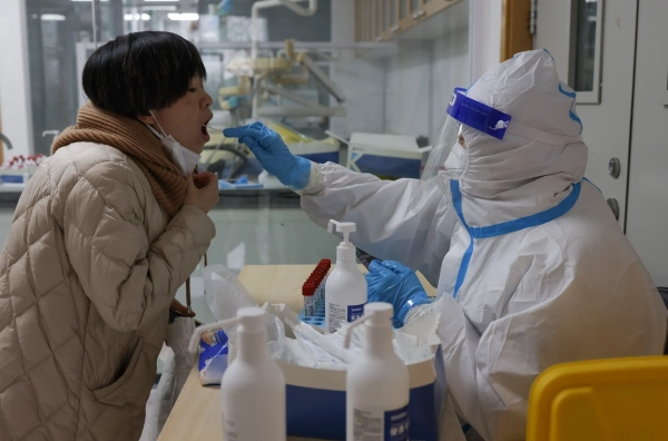 Працівник у білому захисному костюмі, рукавичках, капюшоні та масці тягнеться через стіл, щоб взяти мазок з відкритого рота пацієнта.